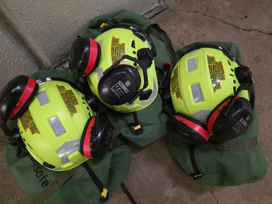 Three hi-vis yellow Sasquatch Pest Control safety helmets with red-and-black ear defenders, resting on green gear bags