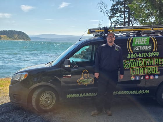 Bryce Carter standing beside the wrapped van on the shore, the water and a forested headland behind him