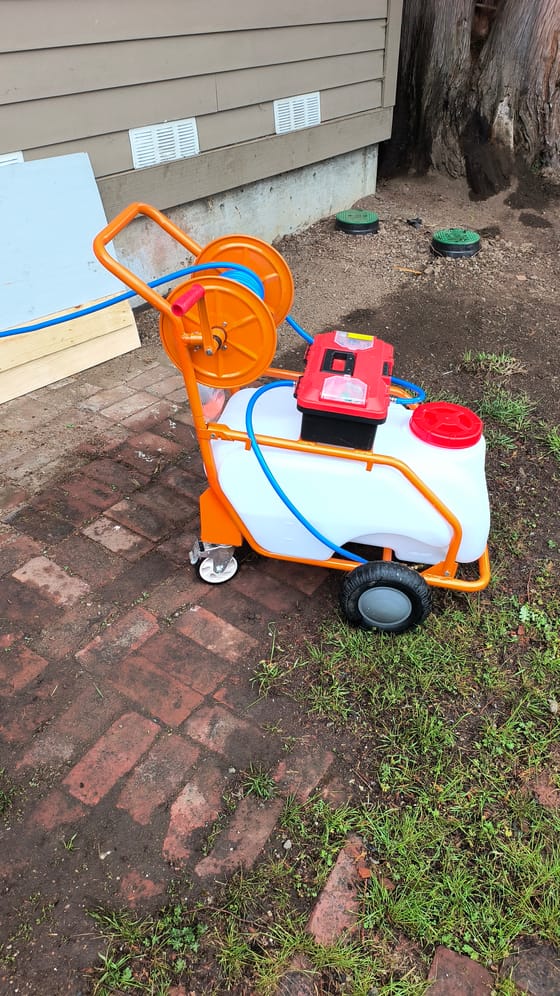 The orange sprayer cart with its white tank and blue hose on a reel, set up beside a house for a crawlspace treatment