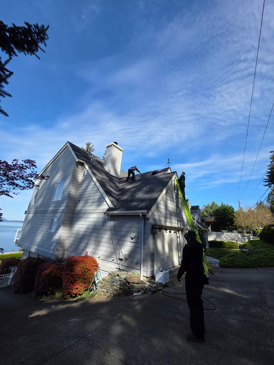 Two technicians in harnesses on the roof of a large gray house above the water, one on the ridge and one on a ladder at the gable, with a third watching from the driveway