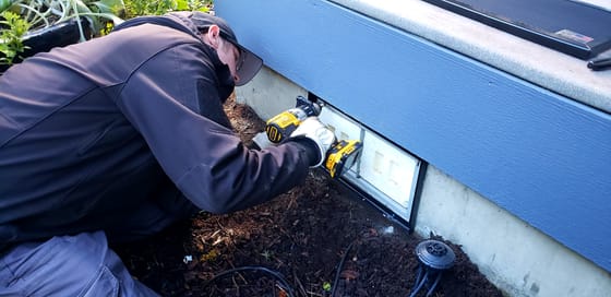 Evan Friese fastening the frame of a steel vent guard over a crawlspace vent with an impact driver