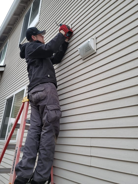 Evan Friese on a stepladder fitting a guarded exhaust vent cover to vinyl siding on a two-story house