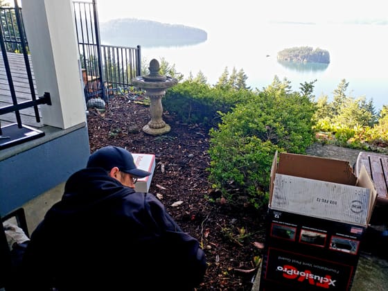 Evan Friese crouched at a foundation vent with the guard box beside him, a bay and two forested islands below the garden