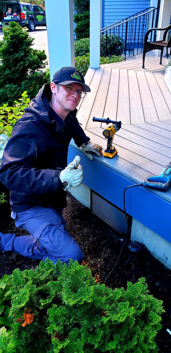 Evan Friese kneeling beside a finished vent guard at the base of a porch, giving a thumbs up, the work truck behind