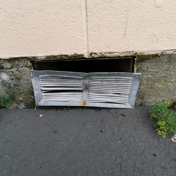 A galvanized louvered foundation vent with bent blades and open corners at the base of a restaurant wall