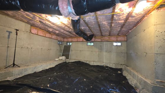 A full-height crawlspace after the job: insulation held up with wire across the joists, new duct, and a black vapor barrier over the floor