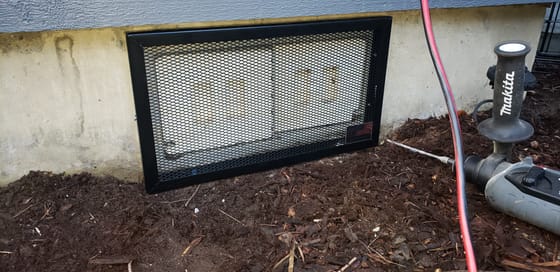 A finished black steel-mesh guard fastened over a crawlspace vent in a concrete foundation, the drill still lying beside it