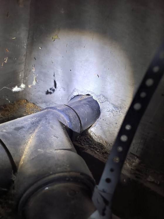A black ABS drain fitting running through a concrete foundation wall, with broken concrete and a gap around the hole