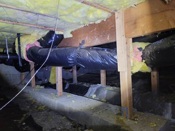 HVAC flex ducts in torn black plastic jackets, fiberglass showing, hung from straps under yellow batts in a crawlspace
