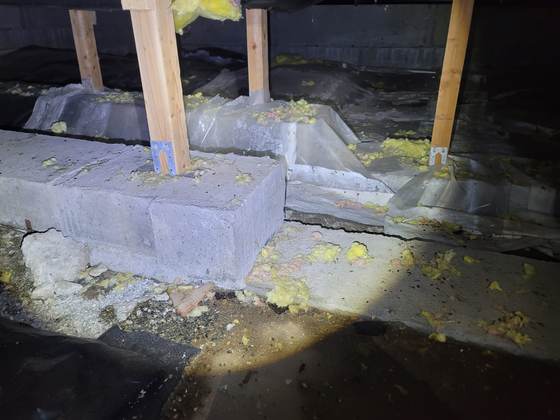 Concrete-block piers and wood posts in a crawlspace, wrapped in plastic sheeting, with shredded yellow fiberglass all around
