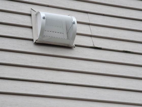 A white guarded cover fitted over a dryer exhaust vent on lap siding, its perforated screen facing out