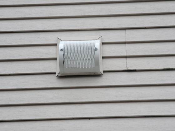 Head-on view of the same white vent guard, its perforated metal screen curved over the opening and screwed to lap siding