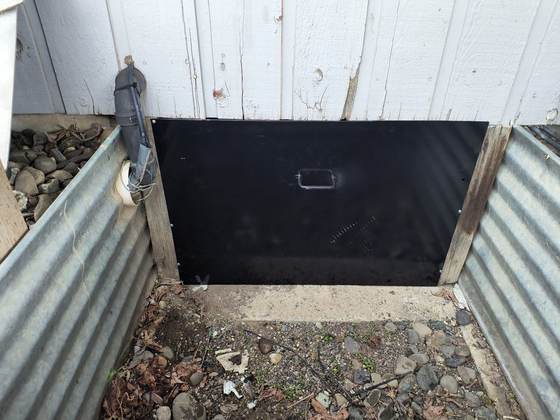 A black powder-coated aluminum crawlspace door set into a metal-lined access well at the base of a white-sided house