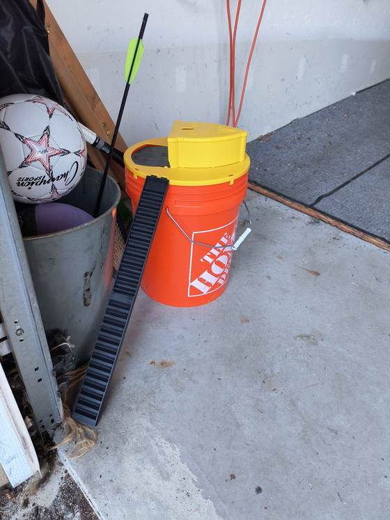 A bucket trap in a garage corner, a ridged plank leaning up to its rim and a lidded plate across the top