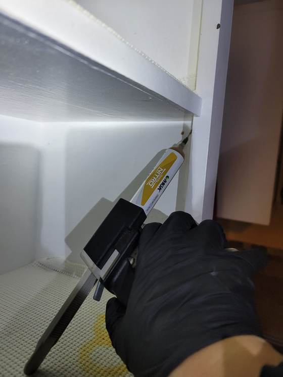 A gloved hand using an applicator gun on the joint under a shelf inside a white cabinet, a textured liner below