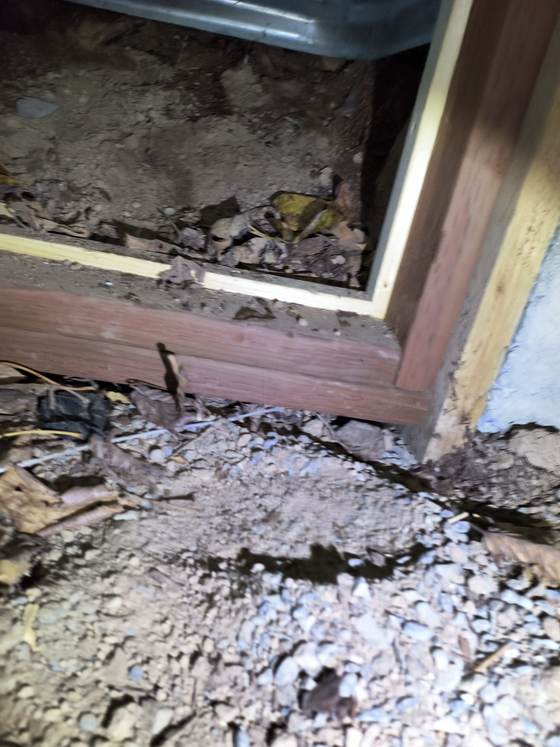 A crawlspace vent opening with gravel and debris in the wood framing