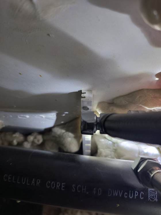 Looking up at black ABS drain pipe, a plastic hanger and a flexible line, with lumps of spray foam packed around them