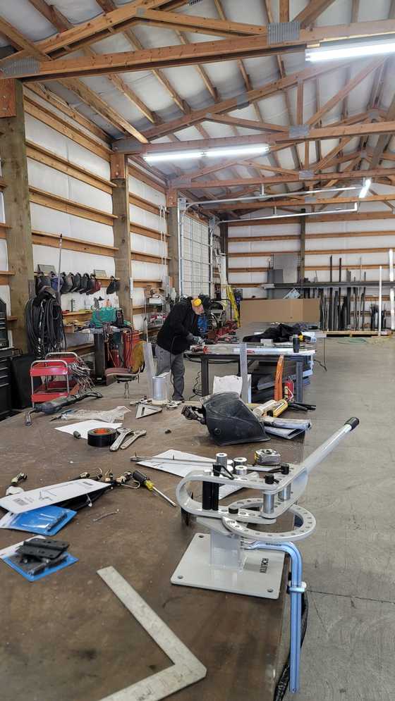 Inside a pole-barn metal fabrication shop: timber trusses, a worker at a bench, a welding helmet and a tube bender up front