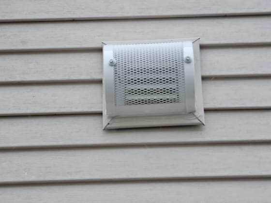 A finished exhaust vent cover with a perforated metal guard, bolted over the vent on beige siding