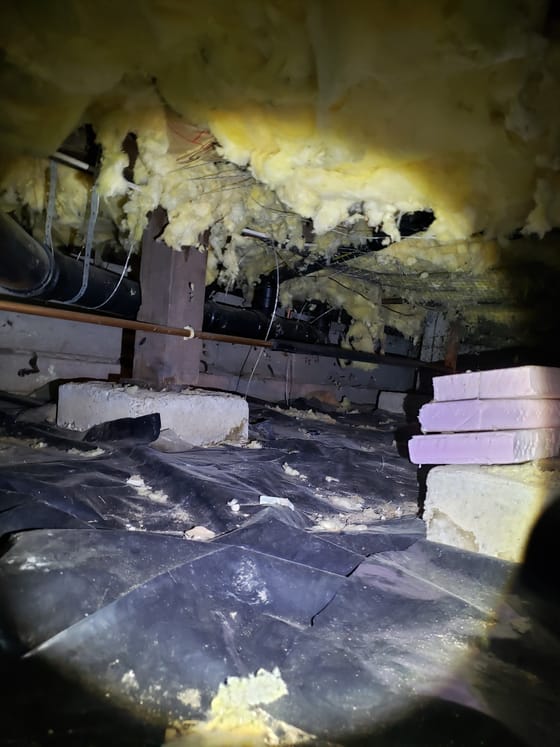 Yellow insulation torn and hanging throughout a crawlspace above piers and a black vapor barrier littered with debris