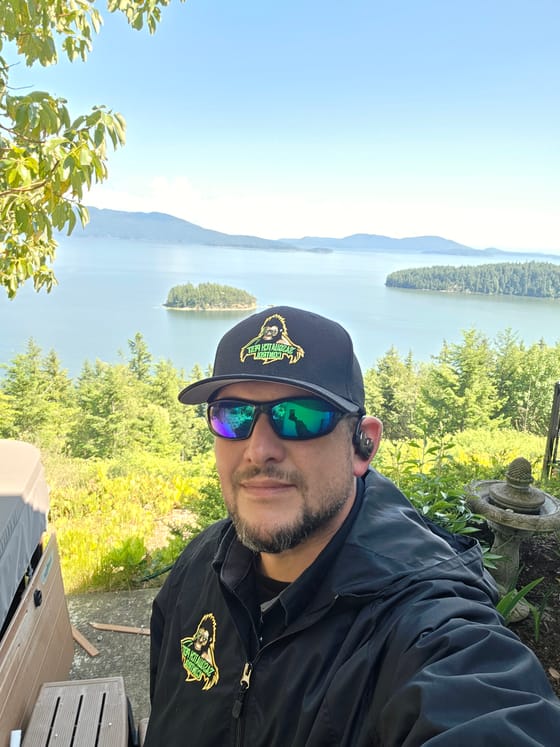 Kristofer Elling in a harness on a roof above Chuckanut Bay, the San Juan Islands behind him