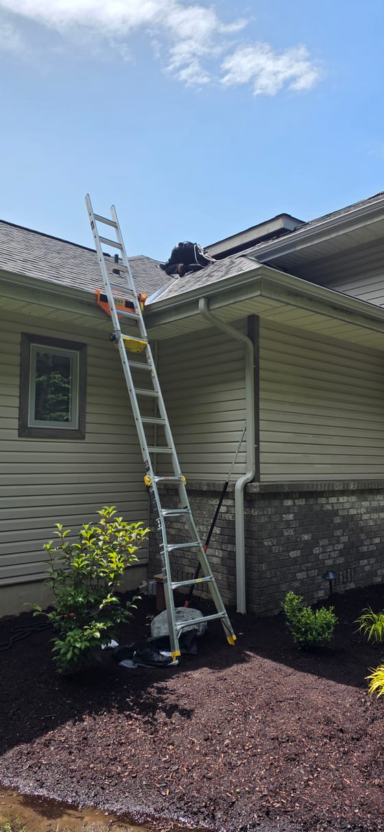 An extension ladder set against a house with a stabilizer bracket at the gutter and a leveler at the base, a technician in a harness working on the roof above