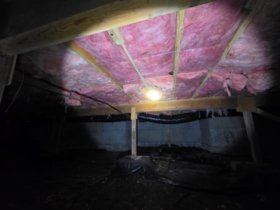 New pink batt insulation held up between the floor joists with wire, over a fresh black vapor barrier, lit by headlamp