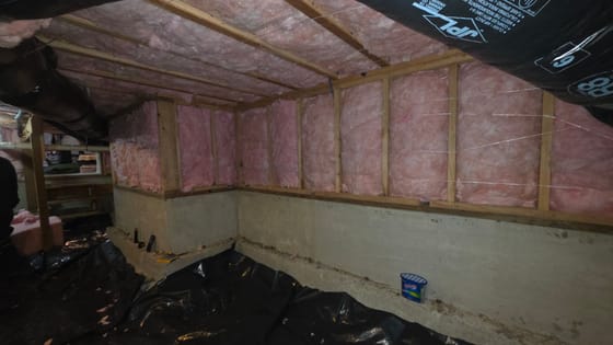 New pink batt insulation filling the framed walls and floor joists of a crawlspace above the concrete foundation, black vapor barrier below