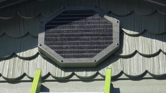 An octagonal gable vent set among scalloped shingles, covered with black wire mesh screwed down at each corner