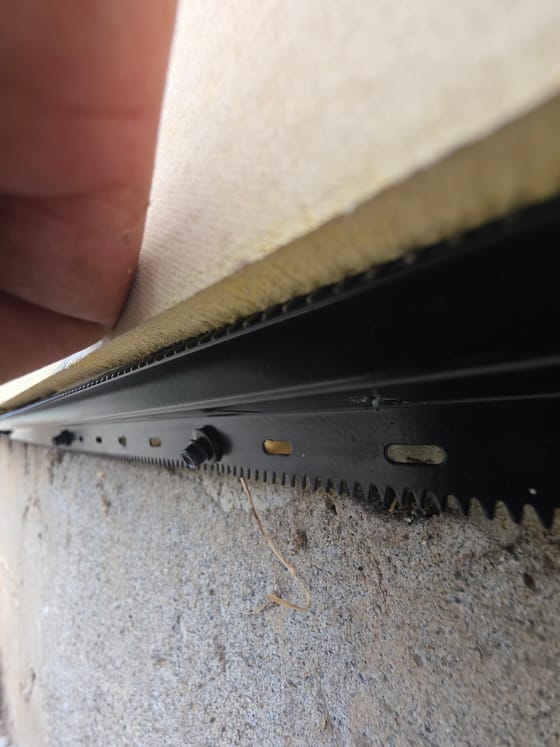 The bottom edge of the siding lifted by hand to show a black toothed rodent shield fastened beneath it against the concrete foundation