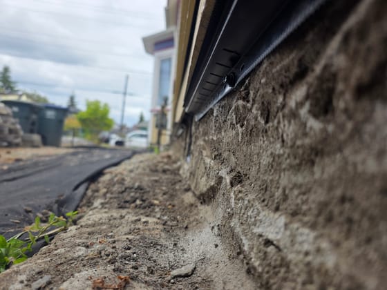 A black metal rodent shield fastened along the bottom of the siding on a house, where it meets a rough concrete foundation, seen low along the wall