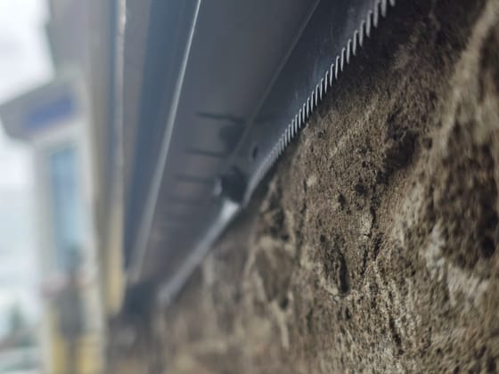 Close-up of the toothed lower edge of a black rodent shield pressed against a rough concrete foundation under the siding