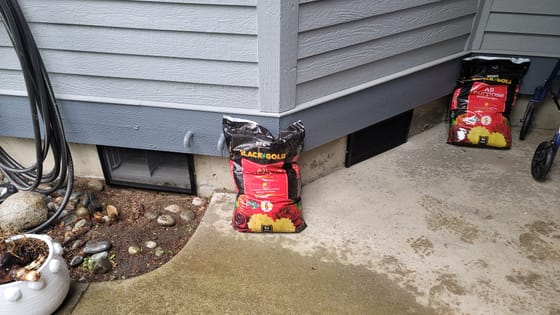 Two black heavy-mesh screens fitted over crawlspace vents along a gray-sided house foundation