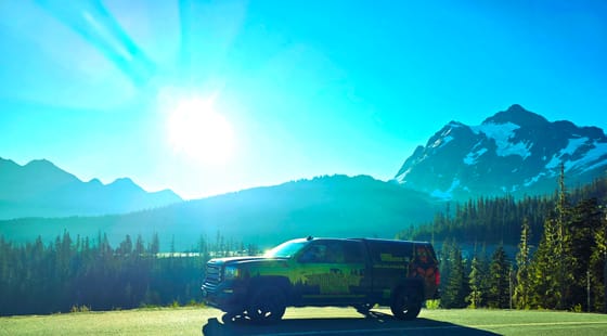 A wrapped service truck parked at a high viewpoint on the Mount Baker Highway, a glaciered peak and forested ridges behind it under a bright sun
