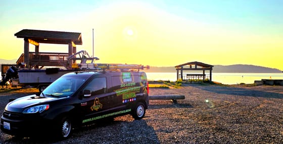 The wrapped van at sunrise on the gravel beside the water on Lummi Shore Road, a boat and timber shelters behind