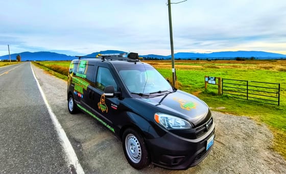 A wrapped service van parked on a farm road shoulder beside green fields and a gate outside Bow, hills on the horizon