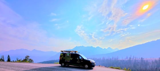 The wrapped van on a mountain road pullout, jagged peaks and forest softened by haze under a hazy sun