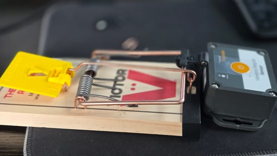 A wooden snap trap fitted with a small black Skyhawk Trapmate vibration sensor at its end, which reports when the trap fires