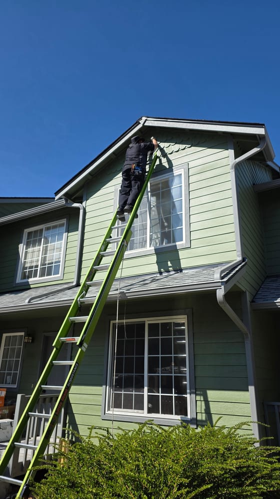 A technician at the top of a tall green extension ladder screening the gable vent of a two-story green townhouse under a clear blue sky
