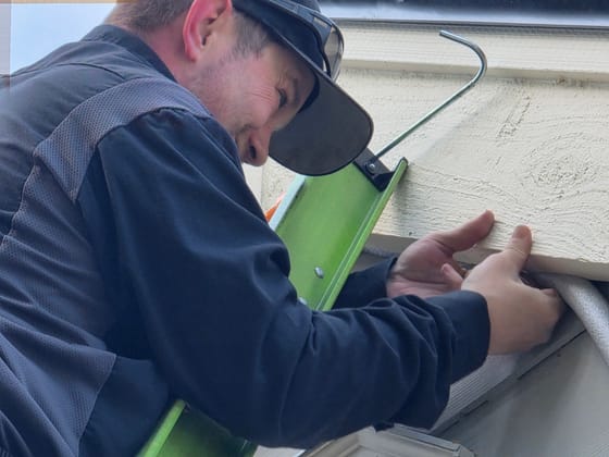A technician in a cap and safety glasses on a green ladder, working a line-set gap under the edge of the siding with both hands
