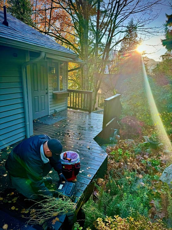 A technician servicing a rodent bait station on a wet deck at a house in Mount Vernon, autumn trees behind