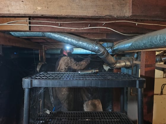 A technician with a headlamp working under metal ductwork and old timber framing in a crawlspace during a powderpost beetle treatment