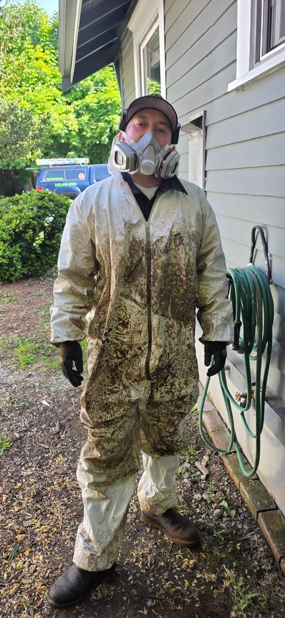 Travis (TJ) Hansen standing beside a house in a mud-covered white protective suit, respirator and gloves after coming out of a crawlspace