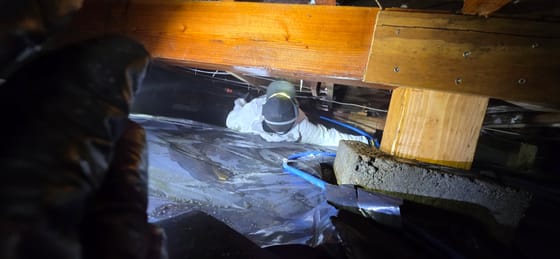 Travis (TJ) Hansen in a white suit and headlamp squeezed under a beam in a very low crawlspace, running a blue spray hose for a powderpost beetle treatment