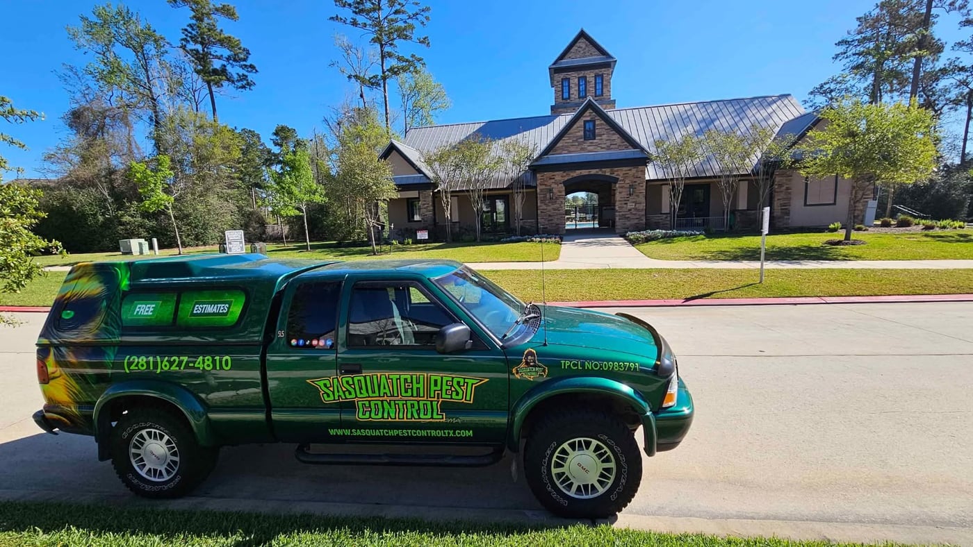 A green wrapped Sasquatch Pest Control pickup parked on a street in The Woodlands, Texas, in front of a stone and metal-roofed community building under tall pines