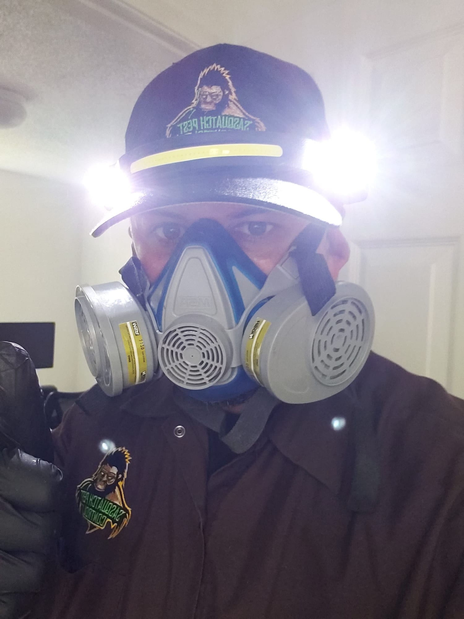 Kristofer Elling in a half-face respirator, cap light and work jacket in a cabin interior, gloved and ready to treat