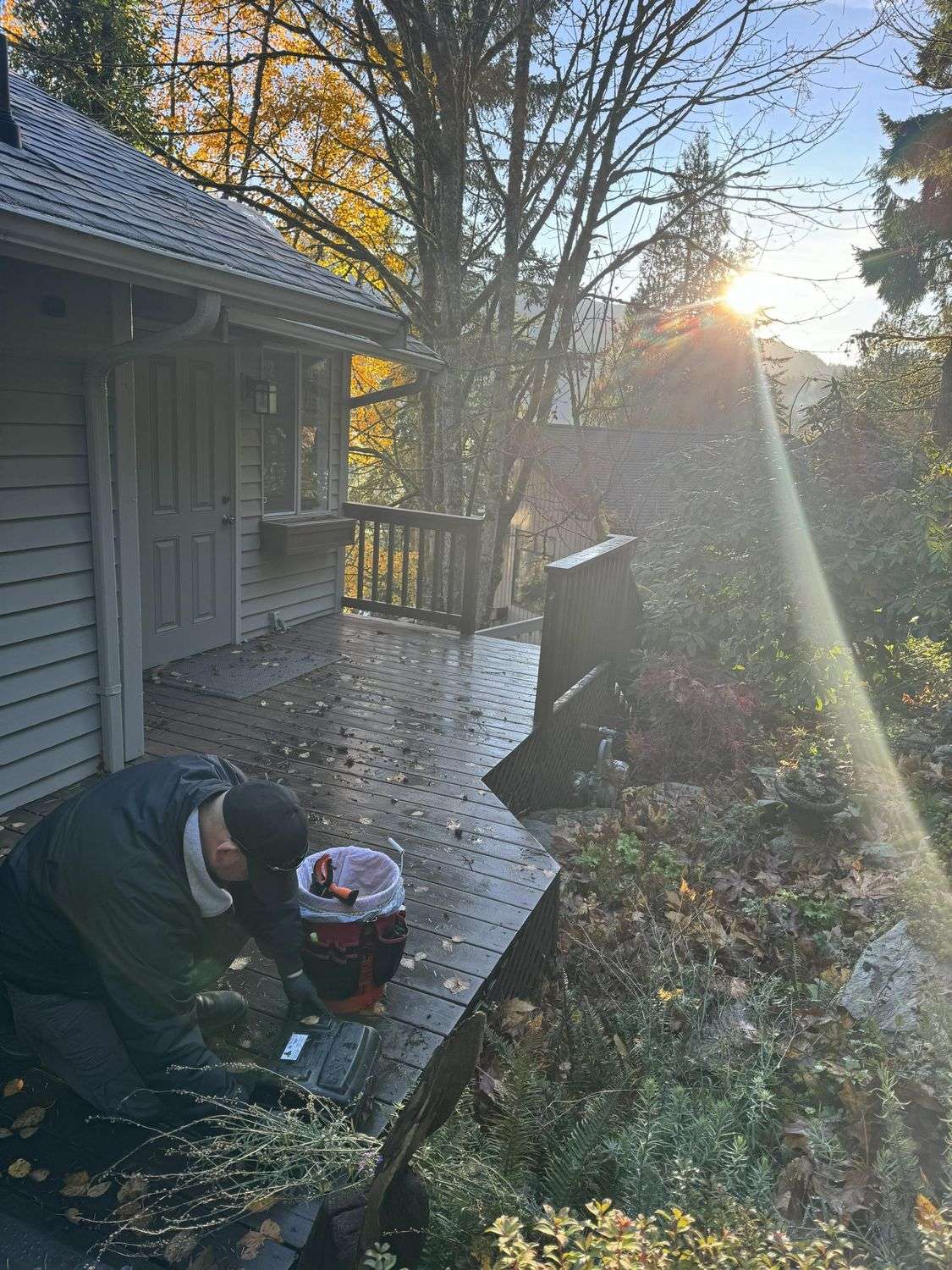 A pest control technician kneeling to work at the base of a house wall beside a deck, low sun coming through the trees behind