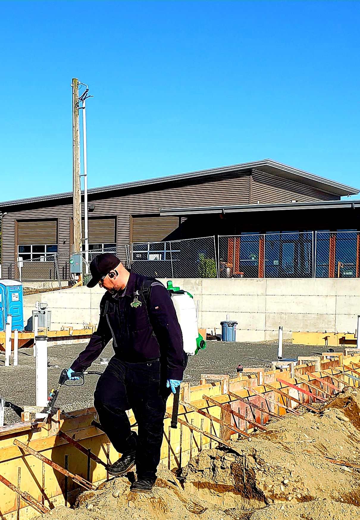 A technician with a backpack sprayer treating soil inside timber foundation forms on a commercial building site under a clear sky