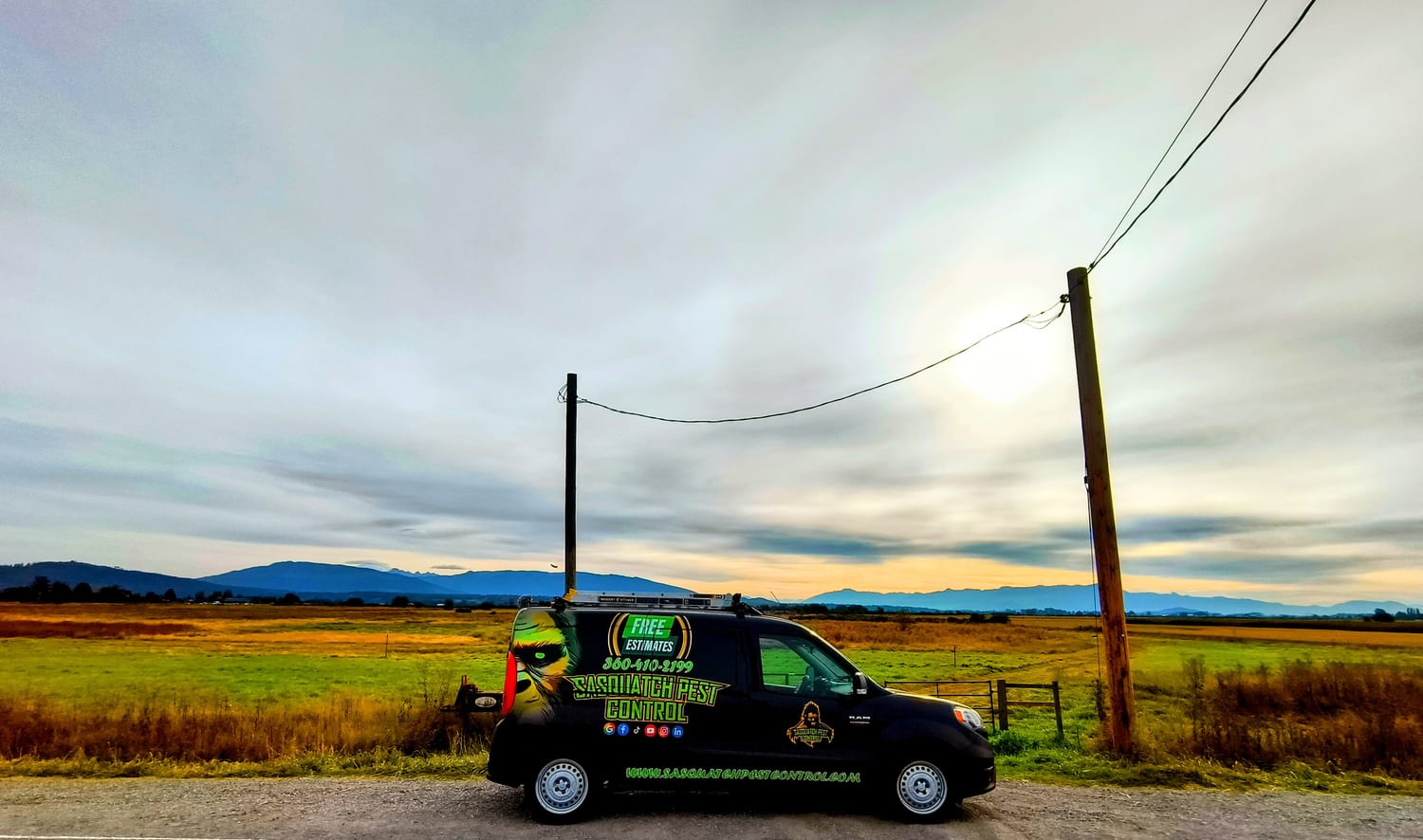 A wrapped Sasquatch Pest Control van parked on a gravel shoulder beside open green and golden fields, with a gate, power poles and a line of hills under a wide cloudy sky