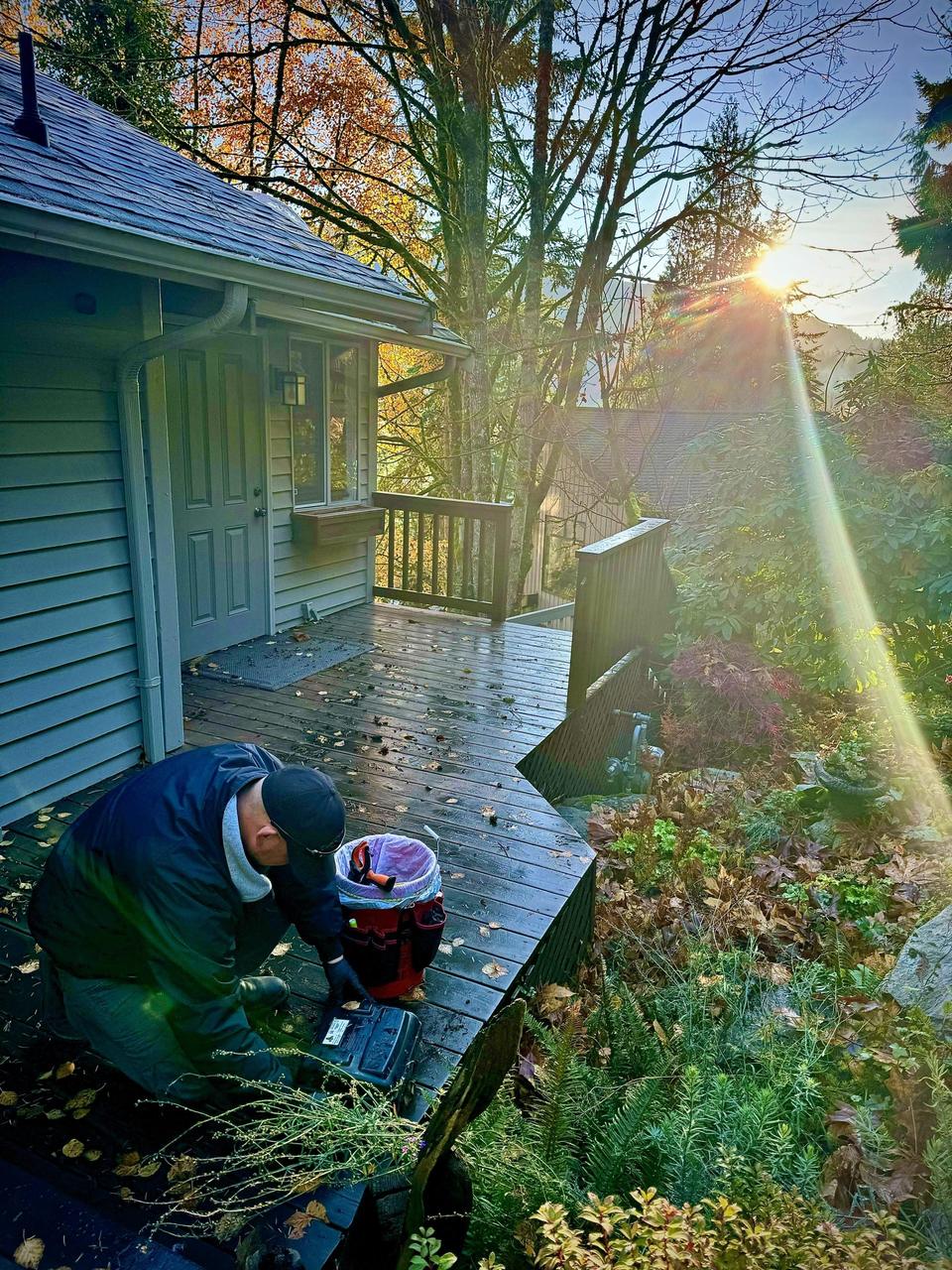 A technician kneeling with equipment beside the deck of a house in low autumn sunlight, with fallen leaves across the boards